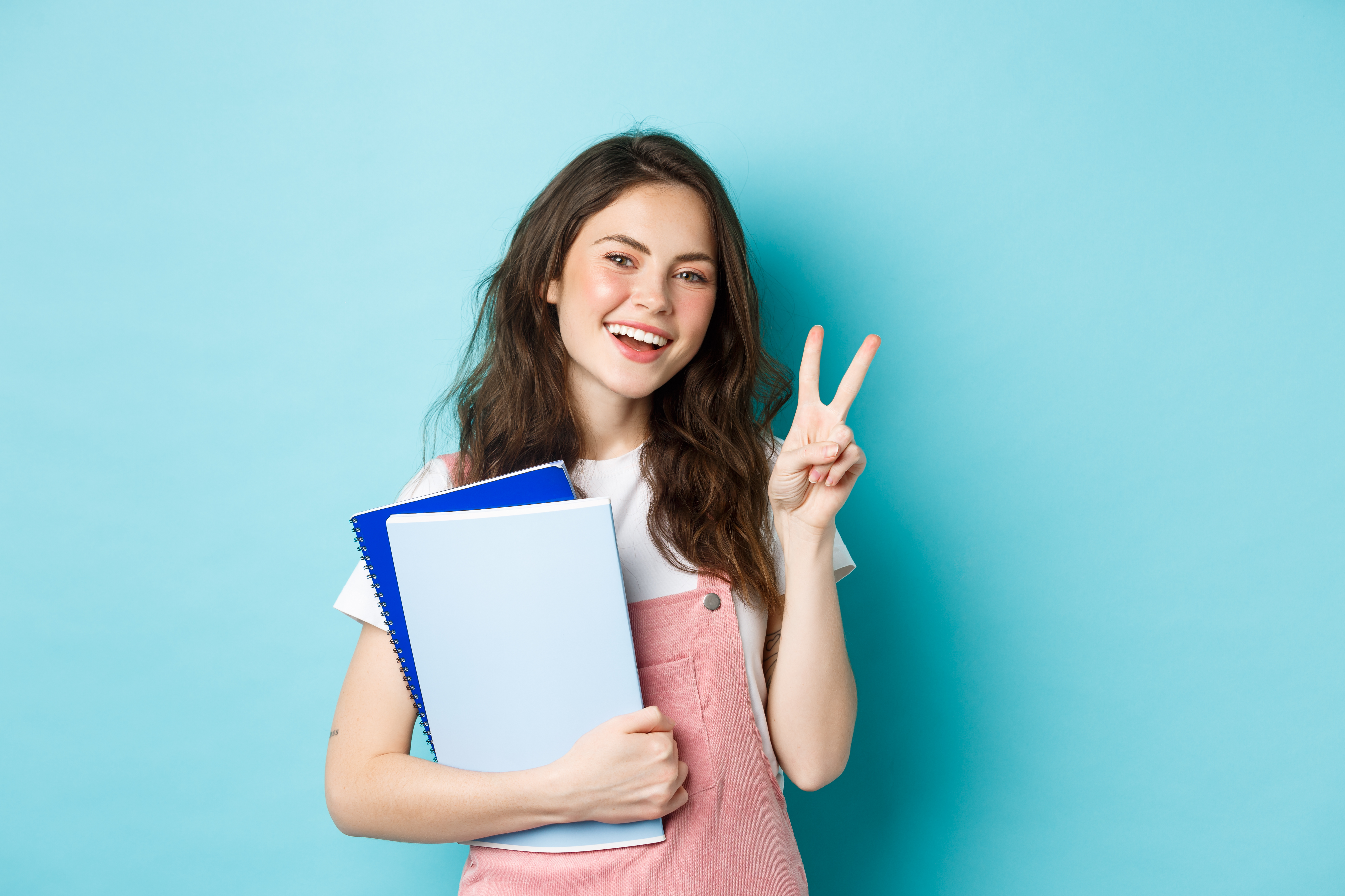 beautiful-female-student-showing-v-sign-smiling-happy-holding-notebooks-with-study-material-attending-courses-standing-blue-background